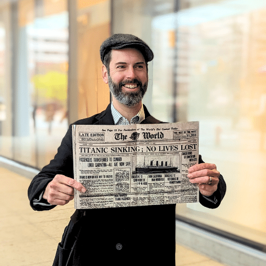 A bearded tour guide with big smile and flat cap holds up a vintage newspaper saying